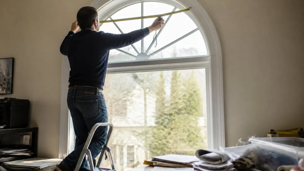 A homeowner on a step ladder carefully measuring the curved top of an arched window with a metal tape measure, notepad and pencil nearby, natural daylight filling the room