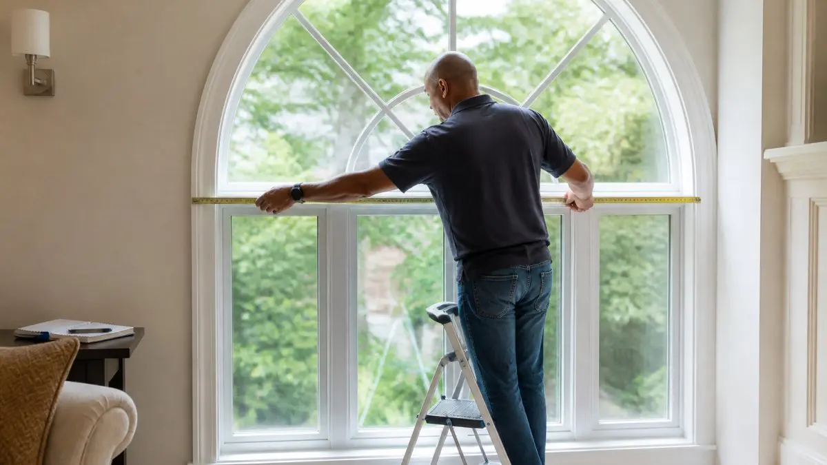 Homeowner on a ladder measuring the base width of large arched windows with a tape measure
